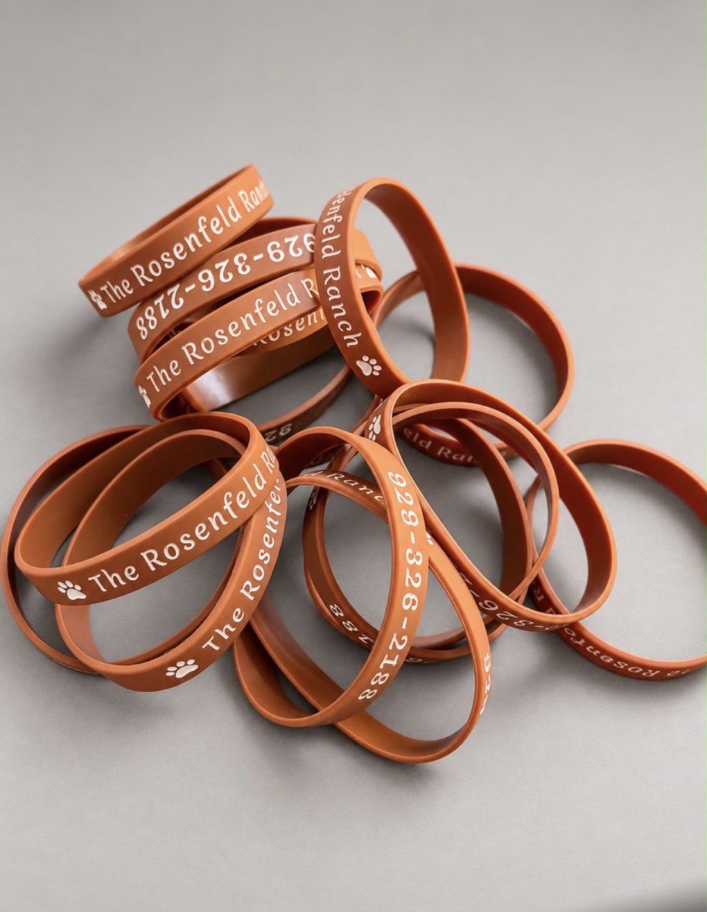 Stack of rust-orange Rosenfeld Ranch silicone bracelets printed with a paw print and (929) 326-2188